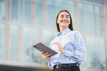 Fototapeta premium Young Professional Woman Smiles While Holding a Tablet Outside a Modern Office Building in Daylight