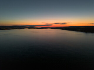 A serene aerial sunset over a calm lake, with subtle reflections on the water and a gradient sky transitioning from deep blue to warm orange hues.
