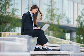 A Woman Working on a Laptop While Sitting on Outdoor Steps in a Modern Urban Area During the Day