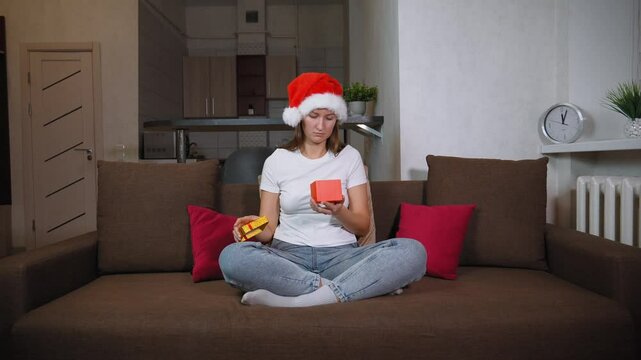 Woman in Santa hat sitting on sofa opening christmas gift. Frustration washes over her face as she opens contents inside, leading to moment of sadness, she is dissatisfied.