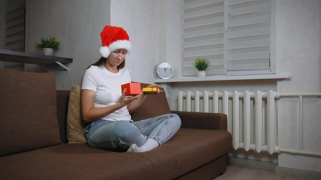 Woman in Santa hat sitting on sofa opening christmas gift. Frustration washes over her face as she opens contents inside, leading to moment of sadness, she is dissatisfied.