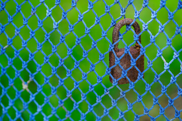 Rusted Padlock Hanging on a Blue Knotted Netting with a Vibrant Green Background Showcasing Contrast Between Security and Nature's Serenity