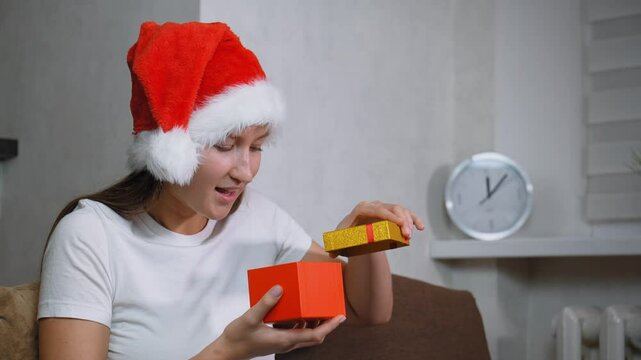 Woman in Santa hat sitting on sofa opening christmas gift. Frustration washes over her face as she opens contents inside, leading to moment of sadness, she is dissatisfied.