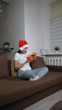 Woman in Santa hat sitting on sofa opening christmas gift. Frustration washes over her face as she opens contents inside, leading to moment of sadness, she is dissatisfied. vertical video