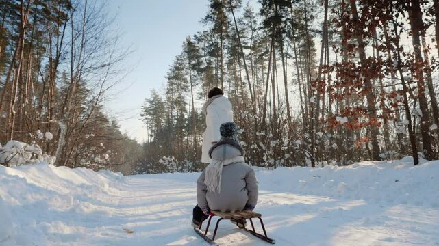 A child sits on a sled while an adult pulls them along a snowy forest path on a bright winter day