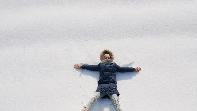 A child in warm winter clothing lies on the snow with arms spread wide, enjoying the winter weather and fresh white snow