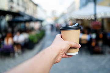 A hand holding a disposable coffee cup with a black lid, extended towards a blurred urban street scene. The focus is on the cup, suggesting an on-the-go lifestyle or a coffee break in a bustling city.