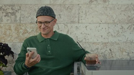 Young Caucasian man sitting at cafe table outdoors, sipping cold coffee drink and scrolling social network page on smartphone
