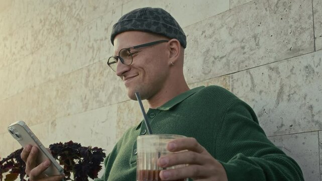 Low angle view of cheerful young Caucasian man sitting at table outdoors sipping bumble coffee and watching videos on smartphone