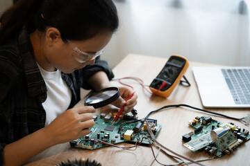 A woman is working on a computer part, looking at it with a magnifying glass. She is wearing safety...