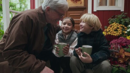 Tracking shot of affectionate grandfather walking along lush garden towards two delighted grandchildren sitting on cozy porch with hot drinks in mugs, wearing casual autumn outfits
