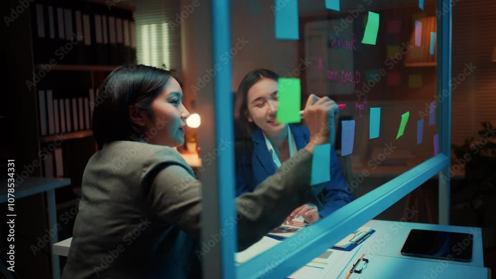 Poster two businesswomen engage in a project discussion, utilizing sticky notes on a glass wall in an offic