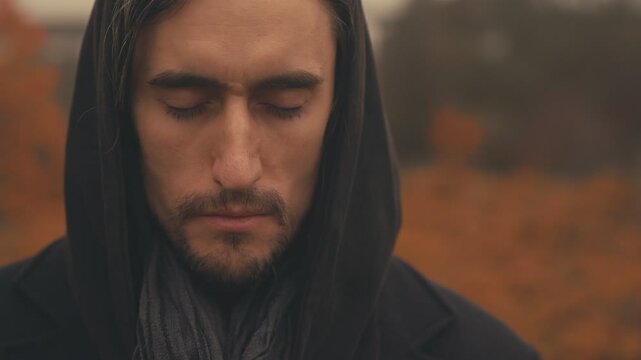 Autumn Sorrow &ndash; Young, attractive European man in black coat with hood, monk-like, surrounded by orange leaves, autumn, bridge in background, close-up, opening eyes, sadness 