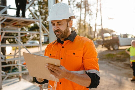 A bearded man wearing a hardhat looking at clipboard on building construction site