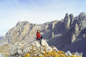 Mountains of Montenegro: a female tourist sits against the backdrop of the peak of Kom Vasojeviсki. Trekking in the Komovi Nature Park: a woman in a bright jacket rests at a viewpoint after climbing