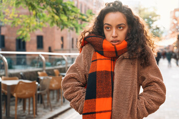 Upset young woman wearing a cozy tan jacket and orange plaid scarf stands in a vibrant urban street...