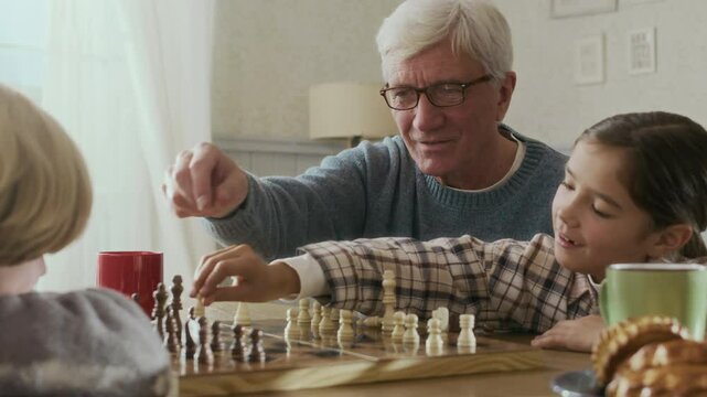 Chest up of gray-haired elderly man in glasses playing game of chess with his two smart grandchildren at wooden table in cozy domestic atmosphere at daytime