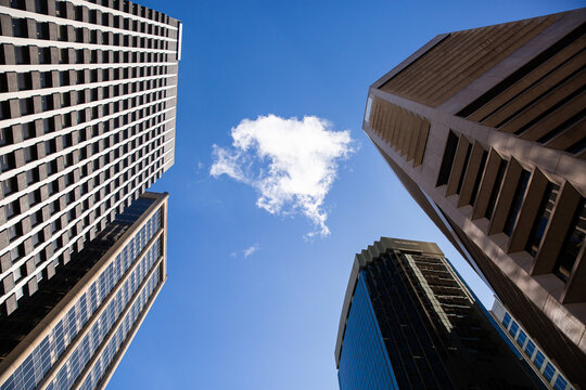 looking up at a single cloud in an otherwise blue sky surrounded by tall city buildings