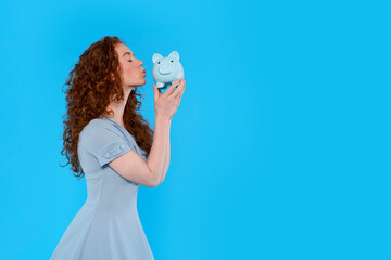 Woman in a light dress kisses a blue piggy bank against a bright blue background