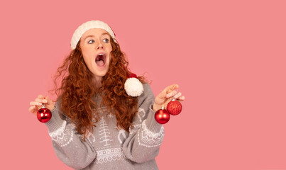 Young woman wearing a Christmas sweater and Santa hat holds festive ornaments against a pink background