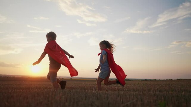 children in superhero costumes. concept of a happy childhood and family for a child. a boy and a girl with red capes and blue T-shirts are running across a field, lifestyle sunset in the background