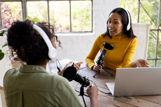 Two multiracial female friends recording podcast at home, engaging in lively conversation - Powered by Adobe