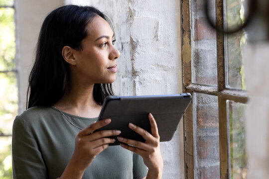 Holding tablet, woman gazing thoughtfully out window in home setting