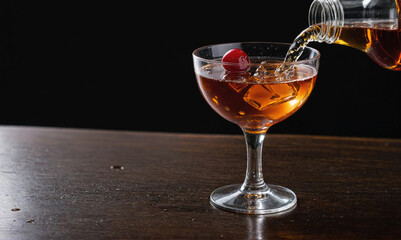 A close-up of a cocktail being poured into a glass with ice and a cherry on a wooden table