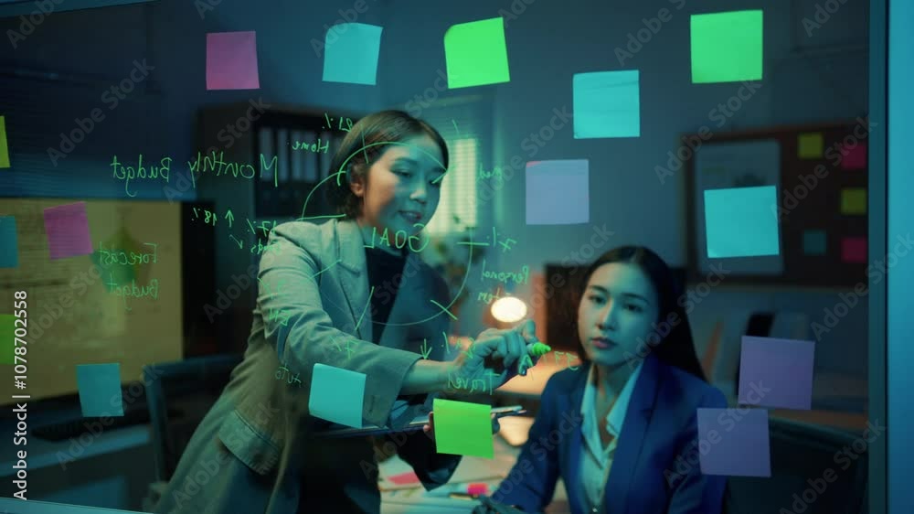 Poster two businesswomen having a discussion about work in the office at night. they are using a glass wall