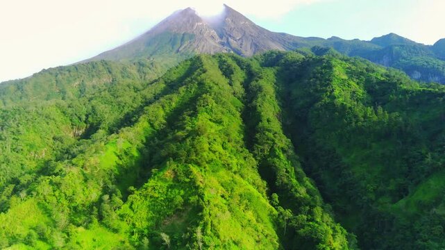 An aerial view of Mount Merapi, Indonesia's most active volcano. Its symmetrical cone is topped by a plume of smoke, contrasting with the lush green slopes and the clear blue sky.