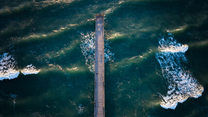 Aerial view looking down on a pier extending into the ocean at Wrightsville Beach, NC, surrounded by dark, swirling waves under the sunlight.