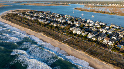 Wide aerial view of Wrightsville Beach, NC, showing a line of beach houses, the ocean, and an extensive inlet area in the background under a clear sky.