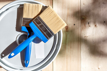 Blue-handled paintbrushes lying on a paint can lid, emphasizing wood surface protection for a wooden house, against a wooden floor background