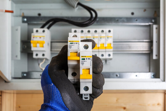 Gloved hand holding two circuit breakers in front of distribution box with multiple switches