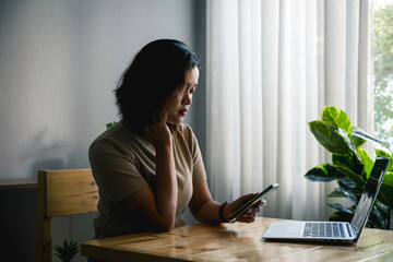 Asian woman with short hair who is stressed while using phone and laptop on table in her home,  unhappy woman