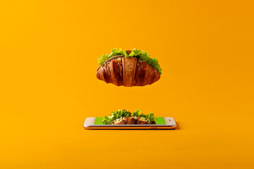A creative concept of online food delivery with a floating croissant over a smartphone on an orange...