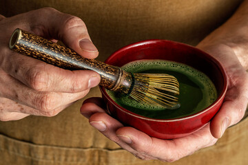 Hands holding a red bowl of matcha tea, whisking the green beverage with a bamboo whisk, capturing...