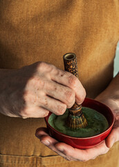 Hands holding a red bowl of matcha tea, whisking the green beverage with a bamboo whisk, capturing...