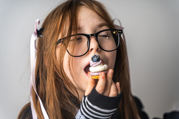 A pretty little girl, a pre-teenager holding a cake in her hands and looking at it smiling, she is dressed in black and white clothes and black eye glasses on a gray background. High quality photo