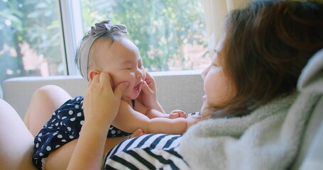 Joyful Mother Holding Smiling Baby by Window with Greenery in Background, mother and her baby enjoying quality time together on a sofa in Cozy Home Environment