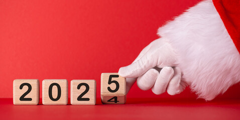 A gloved hand in a Santa suit changes  wooden blocks from 2024 to the year 2025 against a bright red background, symbolizing the New Year countdown and holiday excitement