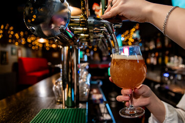 A bartender pours beer from a tap into a glass at a bar, with blurred lights and bar interior in the background. The scene captures the essence of nightlife and social gatherings