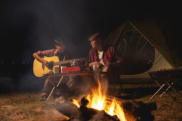 Two teenage friends are having fun playing guitars in front of their tent while camping in forest with bonfire
