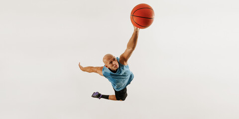 Man dressed in athletic outfit leaps with basketball, preparing for powerful dunk against grey studio background. Concept of sport event, competition, active and healthy lifestyle, game. © Lustre Art Group 