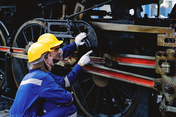 Two men are looking at a train engine. One of them points to the wheel. The other man is wearing a yellow helmet