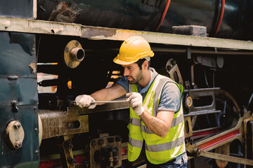 A man in a yellow helmet and safety vest is working on a train engine. He is using a wrench to...
