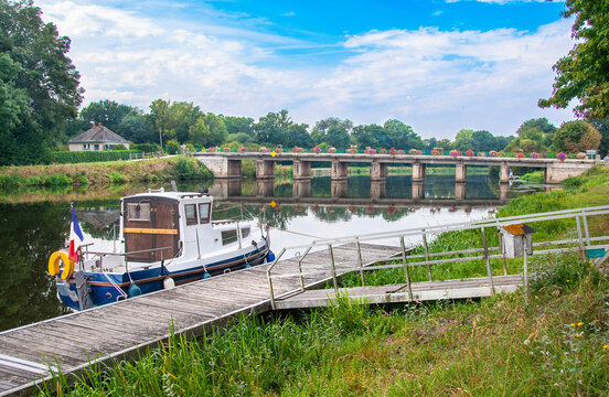 A small, antique French-flagged motorboat is moored in the calm river water close to the reflection of a bridge with flowers near Questembert, France on a summer day.