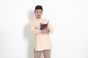 Portrait of excited muslim man in koko shirt with peci writing on his notebook. Happy Asian guy taking notes. Isolated image on white background