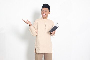 Portrait of excited muslim man in koko shirt with peci reading a notebook. Happy Asian guy pointing to the side with finger. Isolated image on white background