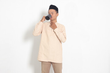 Portrait of attractive Asian muslim man in koko shirt with peci holding a mug, drinking water, quenching thirst after fasting. Isolated image on white background